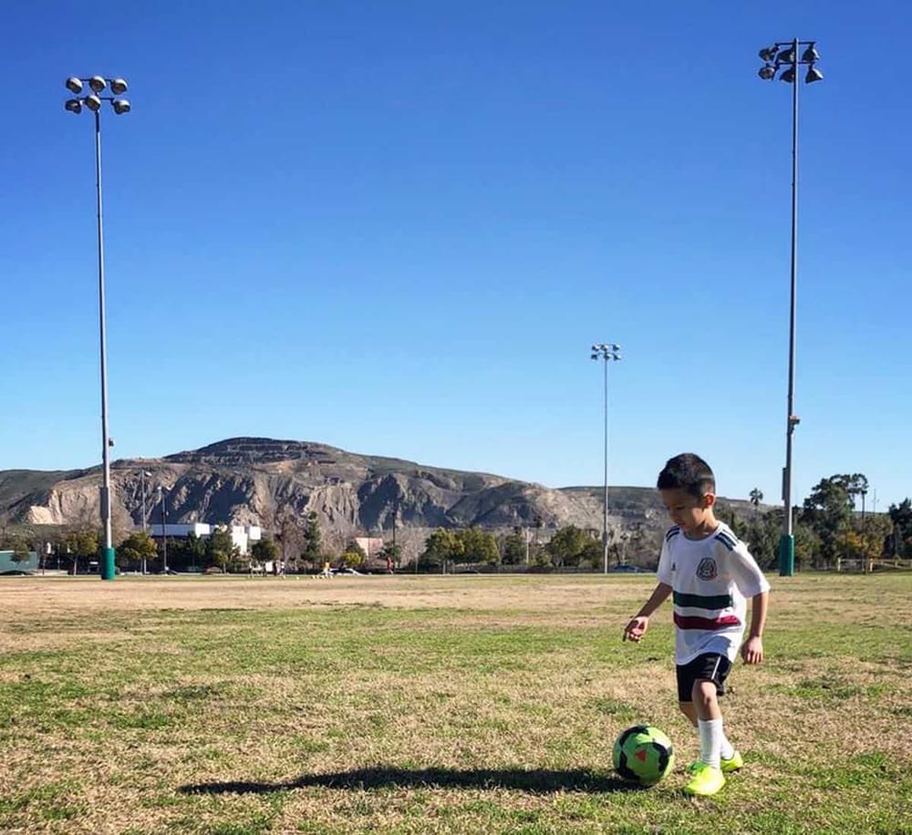 Bright sunny day at the soccer field with young player kicking a ball, scenic mountain backdrop, tall floodlights, QuestForDirections.