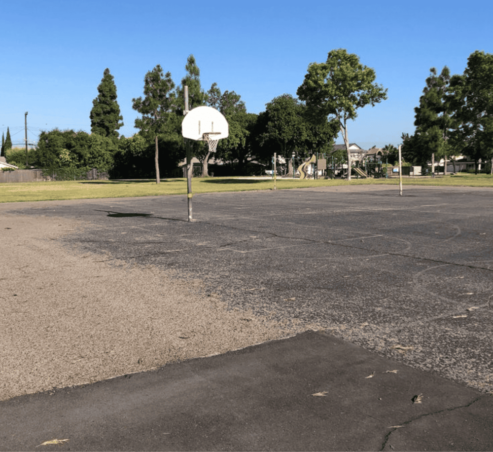 Empty school playground basketball court with trees and playground equipment in the background, ideal for outdoor activities and sports.