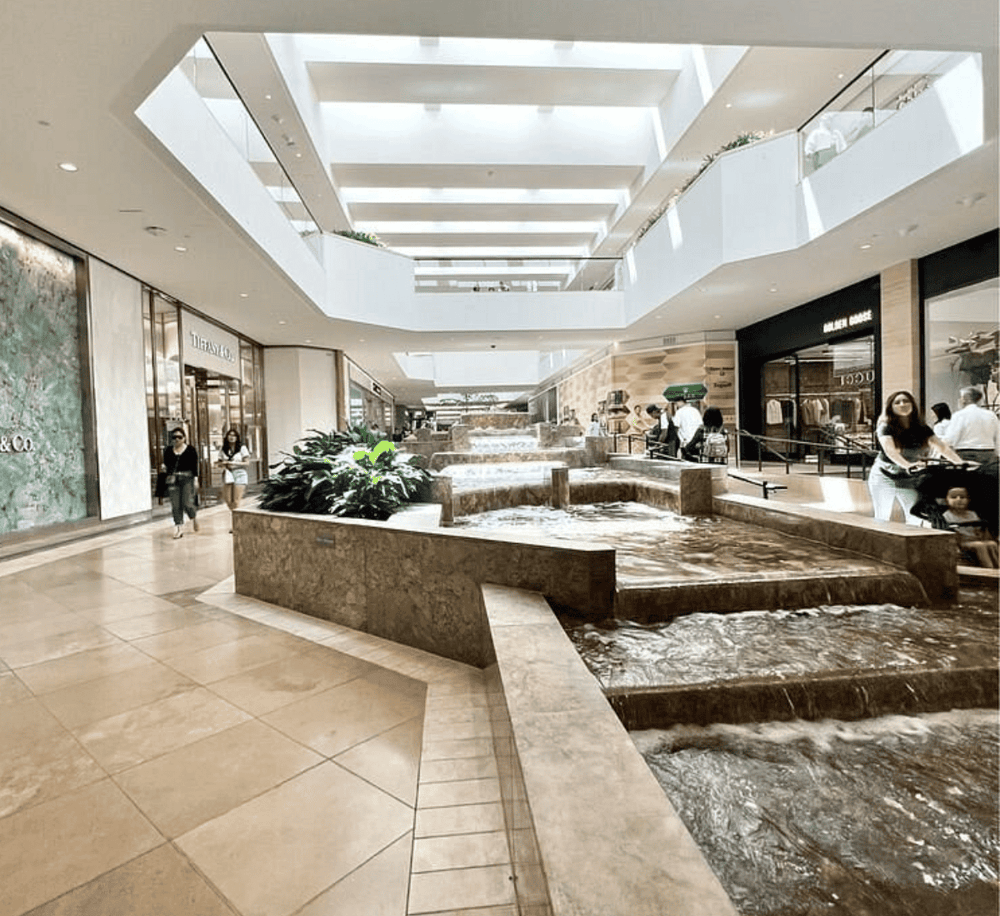 Bright indoor shopping mall with cascading water feature and luxury retail stores.