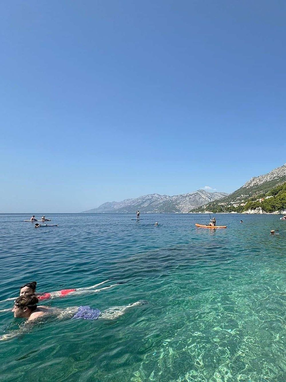 Calm ocean with paddleboarders and swimmers, scenic mountain background, sunny weather, perfect for outdoor water activities.