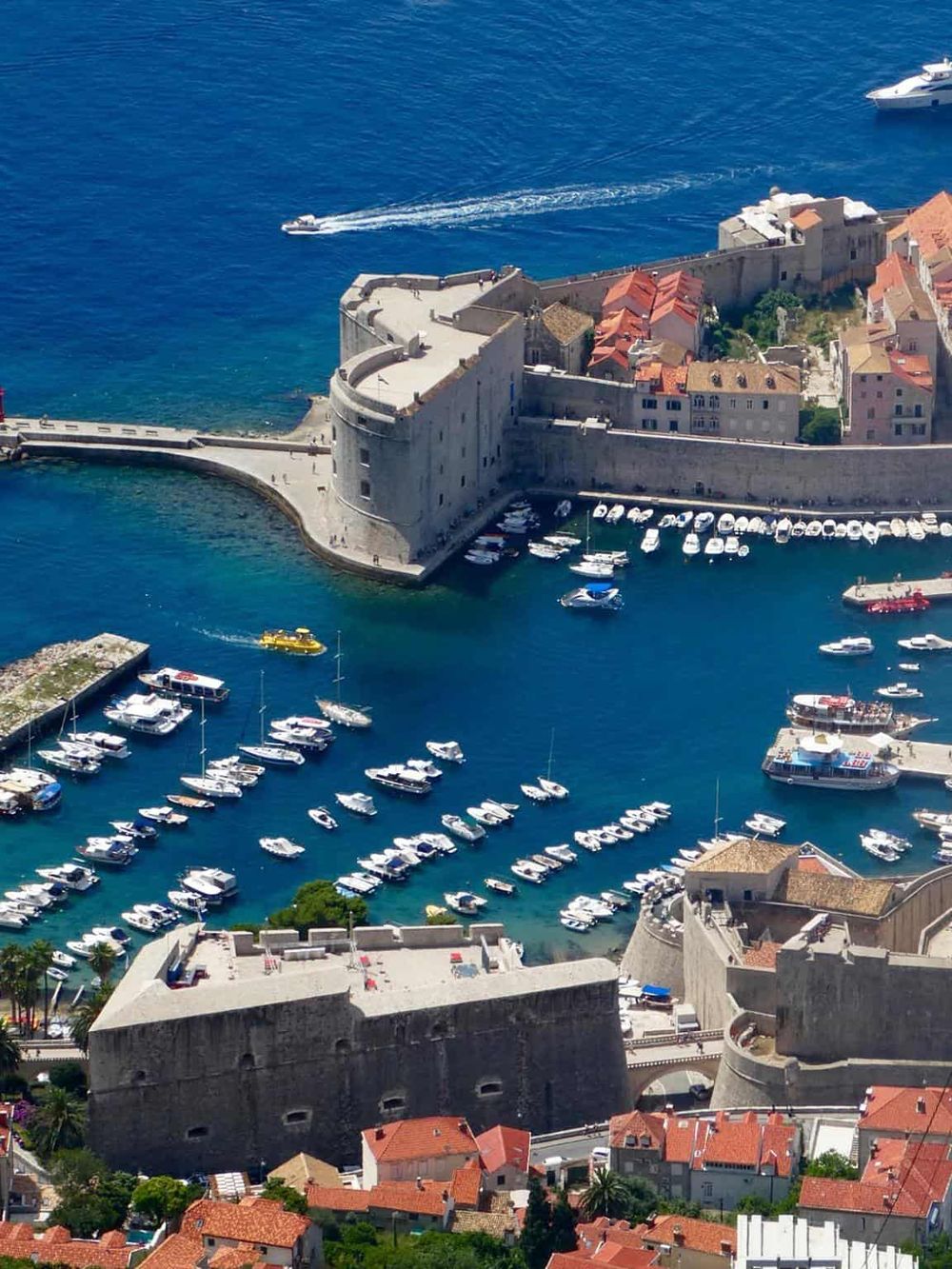Aerial view of Dubrovnik's historic old town and harbor, showcasing medieval city walls, boats, and crystal-clear Adriatic Sea.