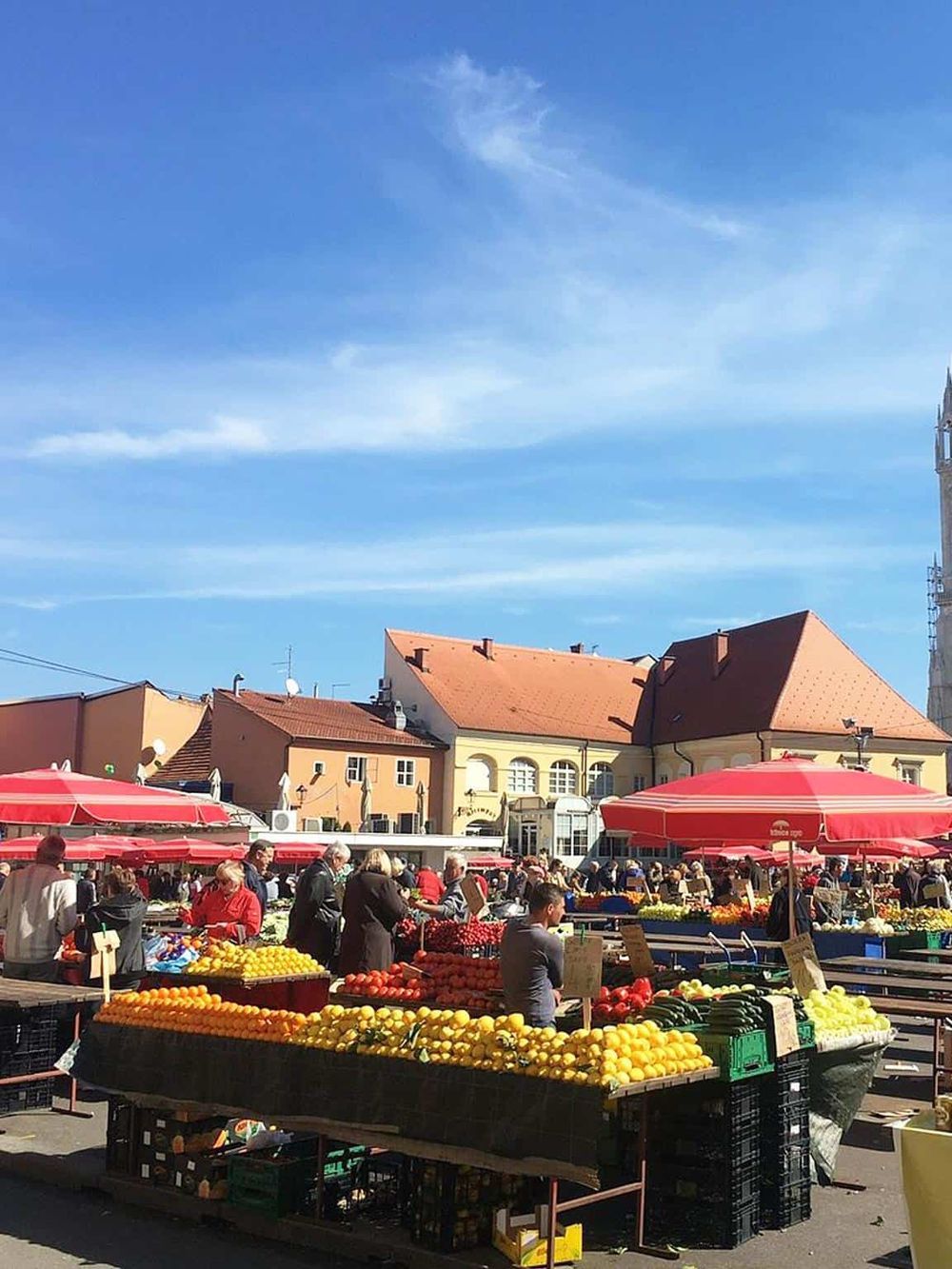 Colorful outdoor farmers market with fresh fruits and vegetables under blue skies, bustling crowd shopping in a European town square.