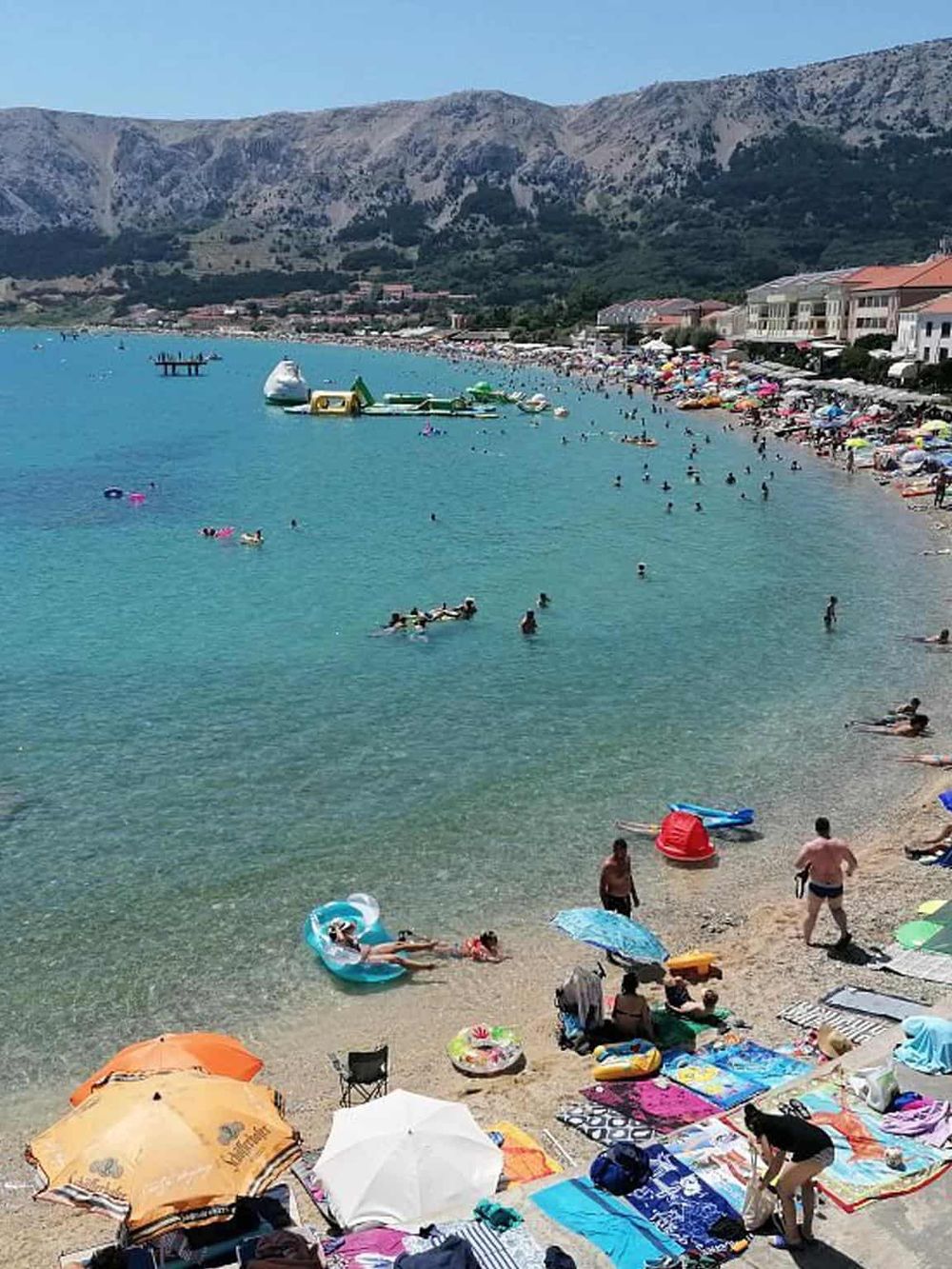 Sunset Beach bustling with swimmers, sunbathers, and colorful umbrellas on a summer day. Turquoise waters and scenic mountain backdrop.