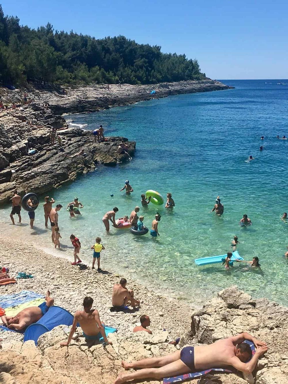 Crowded rocky beach with families swimming and relaxing in clear blue water, lush green trees in background.