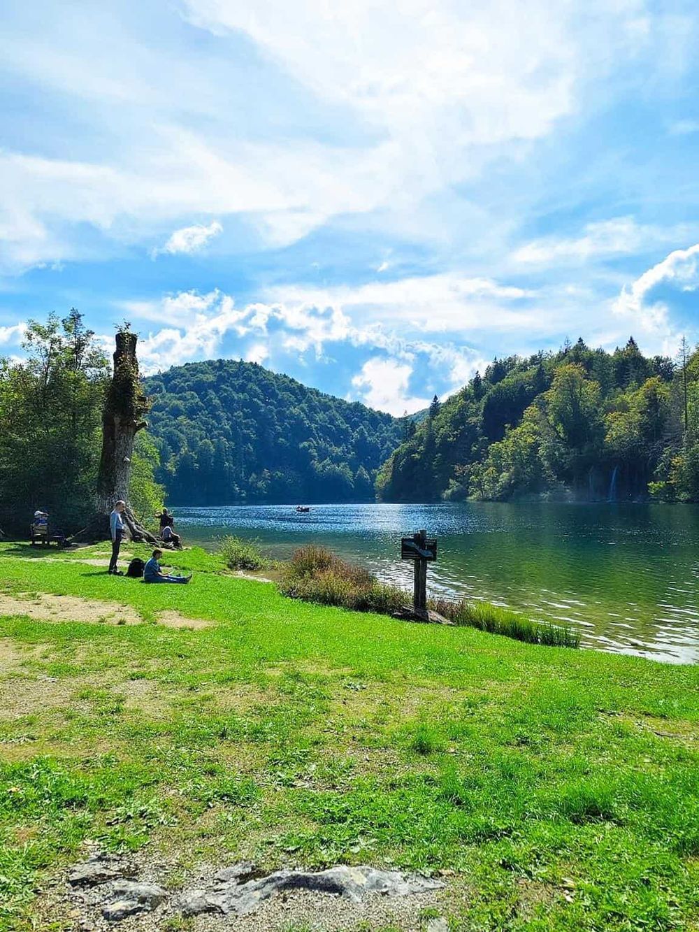 Serene lake surrounded by lush green forest and mountains with people enjoying outdoor activities under a bright blue sky.