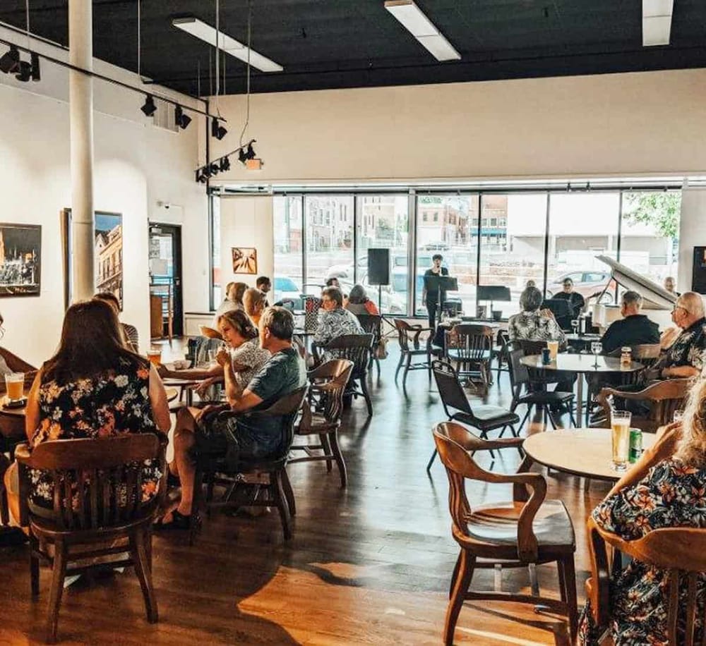 Modern coffee shop interior with customers enjoying drinks and live music performance. Spacious, well-lit, with large windows and cozy atmosphere.