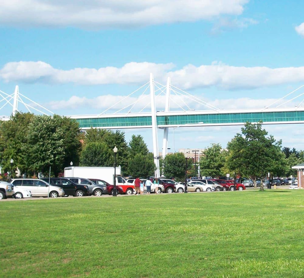 Modern pedestrian bridge with city park, parking lot, and lush trees under blue sky.