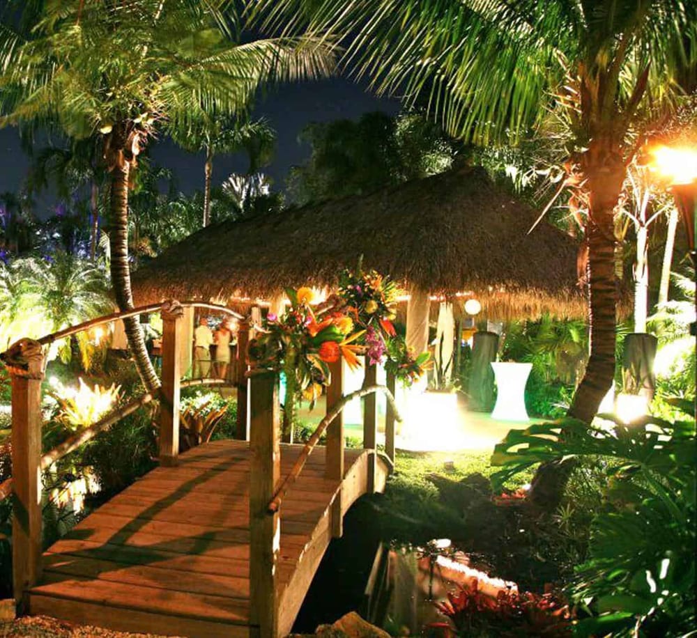 Lush tropical garden at night with bamboo bridge, tiki hut, and vibrant lighting perfect for events.