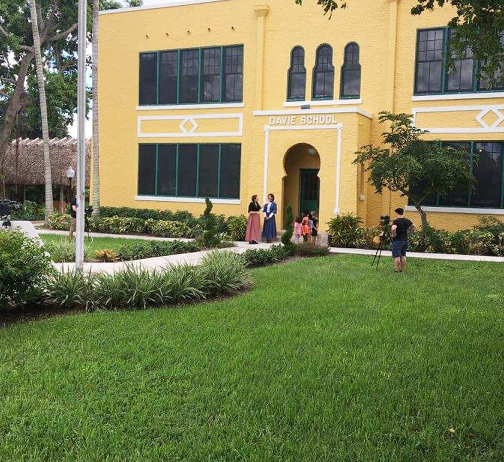 Children and teachers at DAVE SCHOOL entrance in sunny outdoor campus scene.