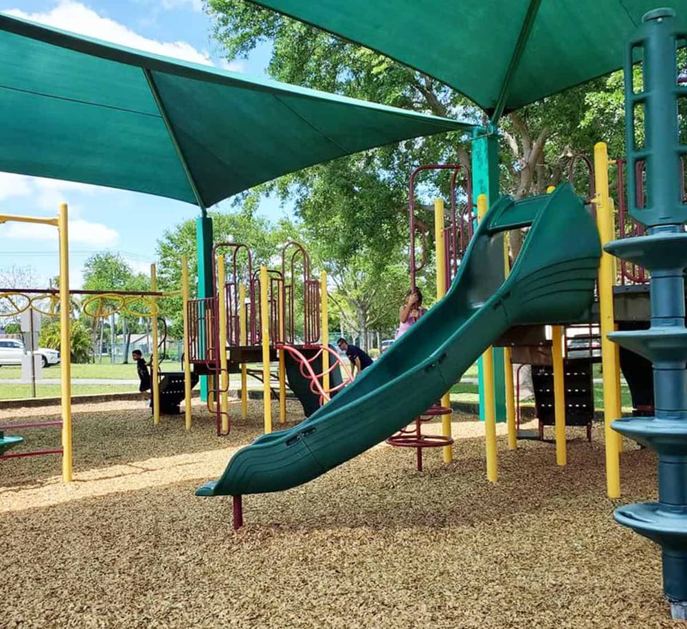 Colorful playground slide and play structures in a sunny park setting with shade sails.