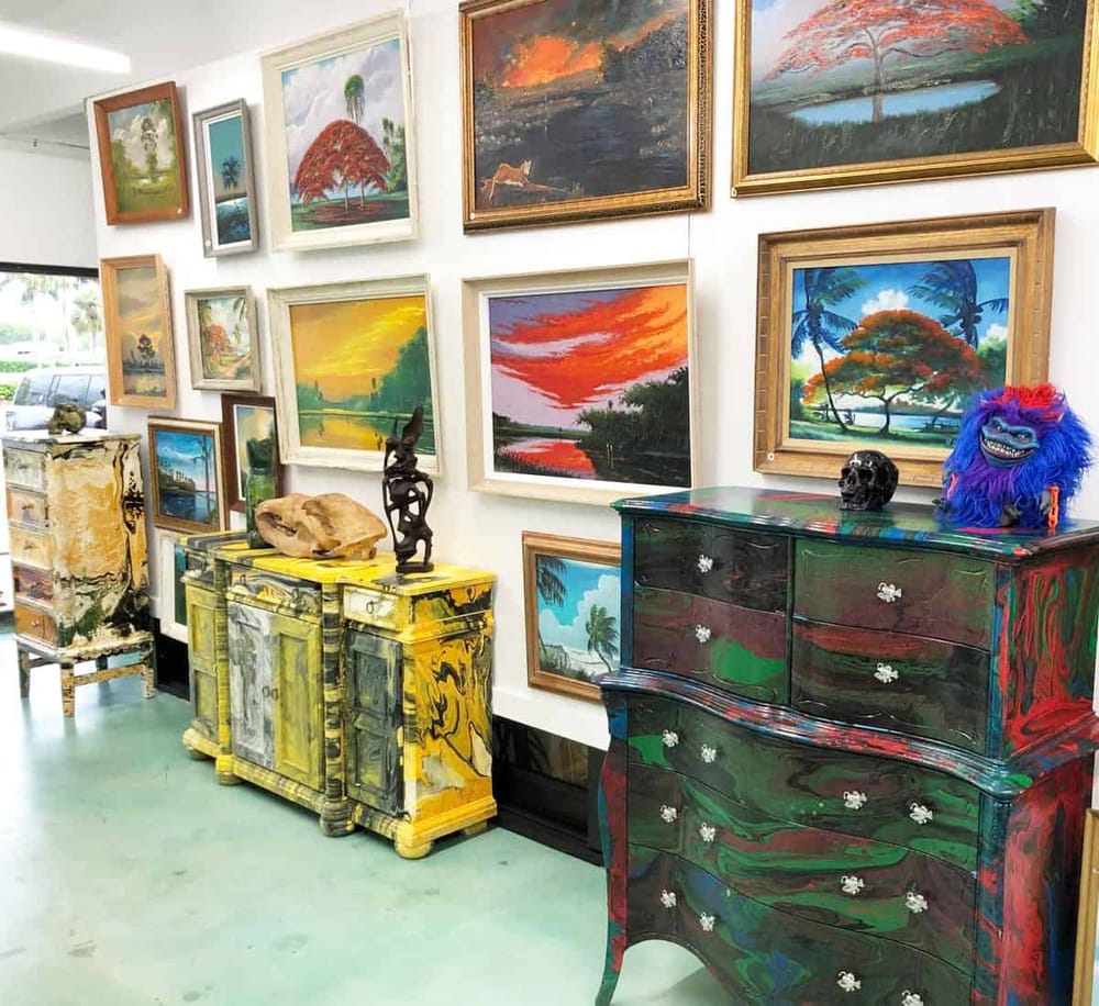 Colorful art gallery with landscape paintings and vibrant painted furniture, showcasing tropical and sunset themes.