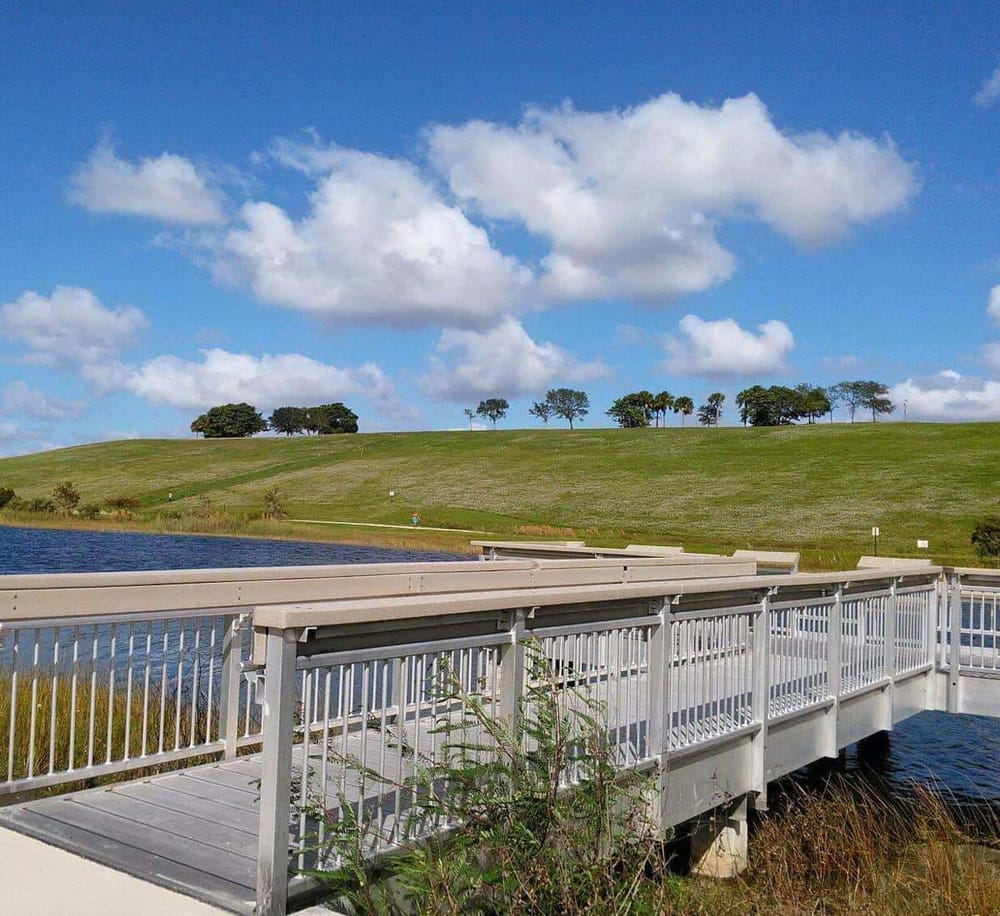 Tranquil outdoor scene with a wooden deck overlooking a pond and green rolling hills under a blue sky with clouds.