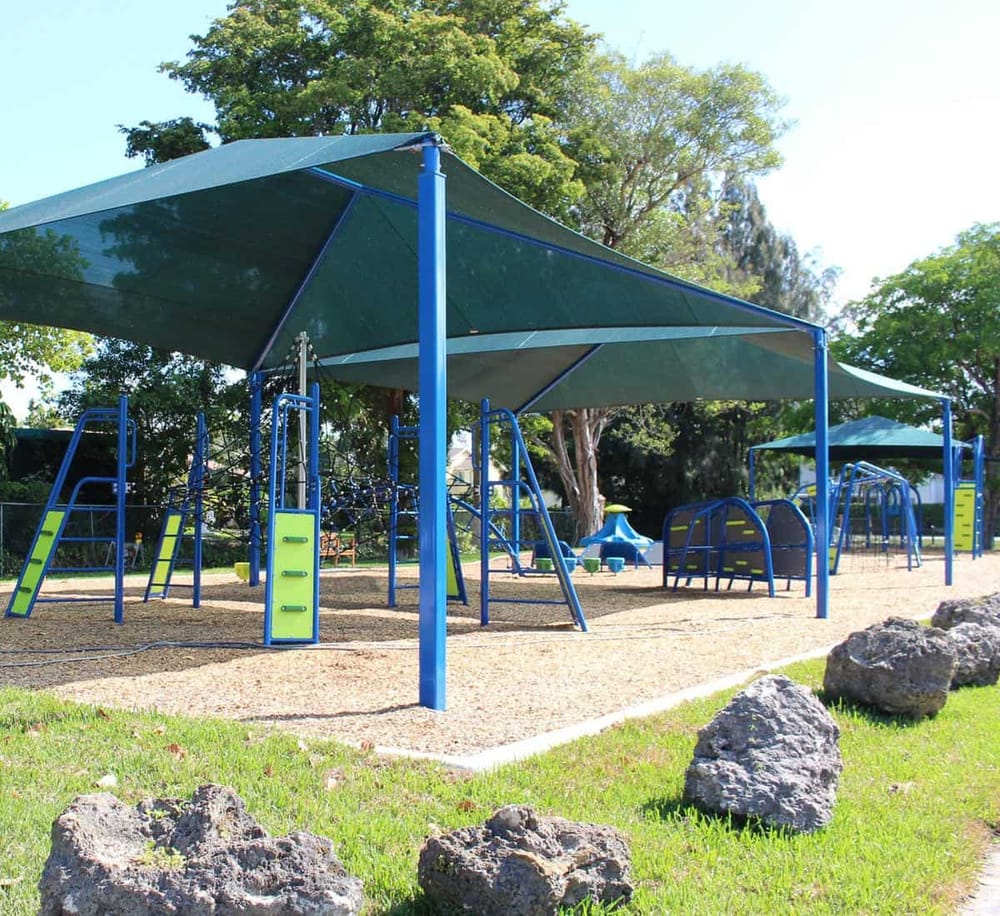 Bright blue playground structure with shade sails in a sunny park with trees.