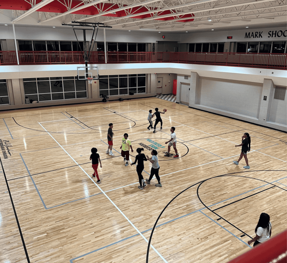 Kids playing basketball in a modern indoor gymnasium with wooden floors and large windows.