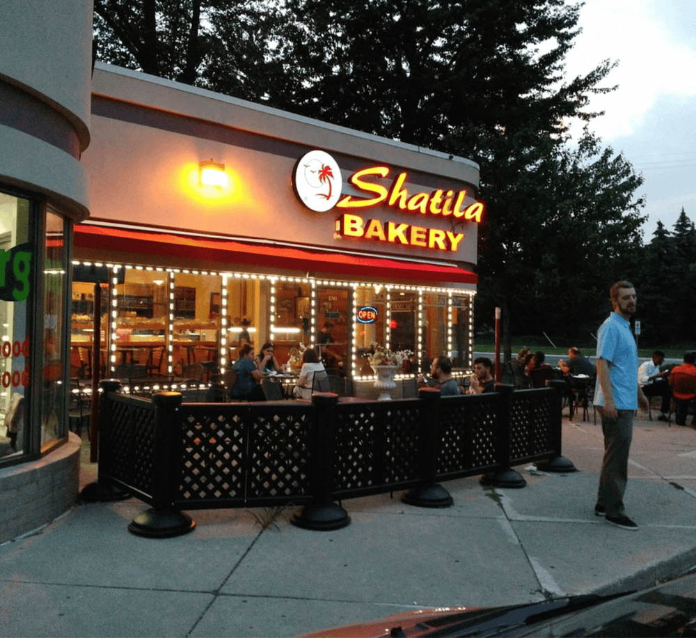 A bright, inviting bakery storefront illuminated with neon signs and outdoor seating at dusk.