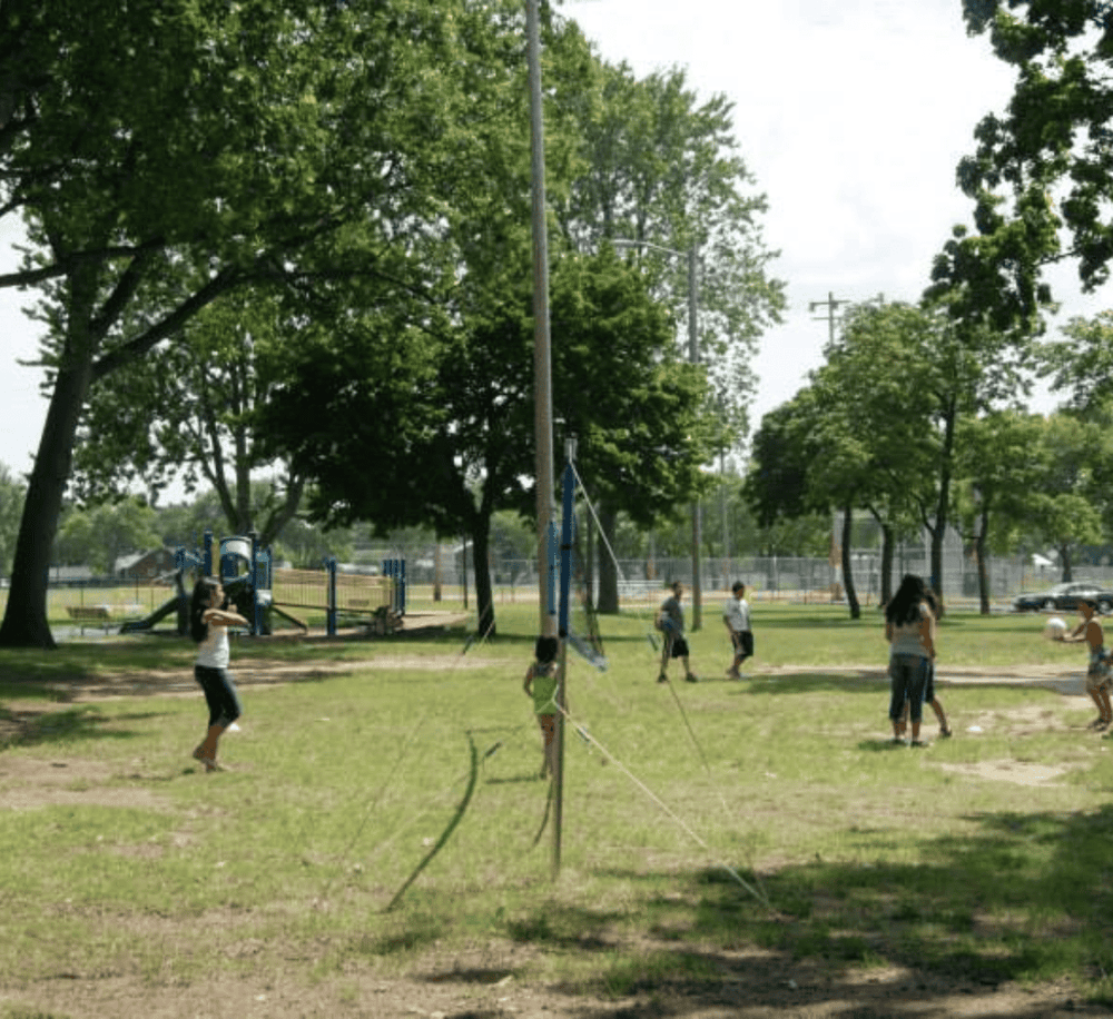 Children playing volleyball in a park with trees and playground equipment.