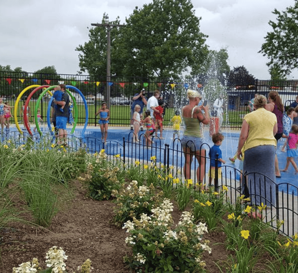 Colorful splash pad with kids playing, families enjoying summer fun, and water features at a community park.
