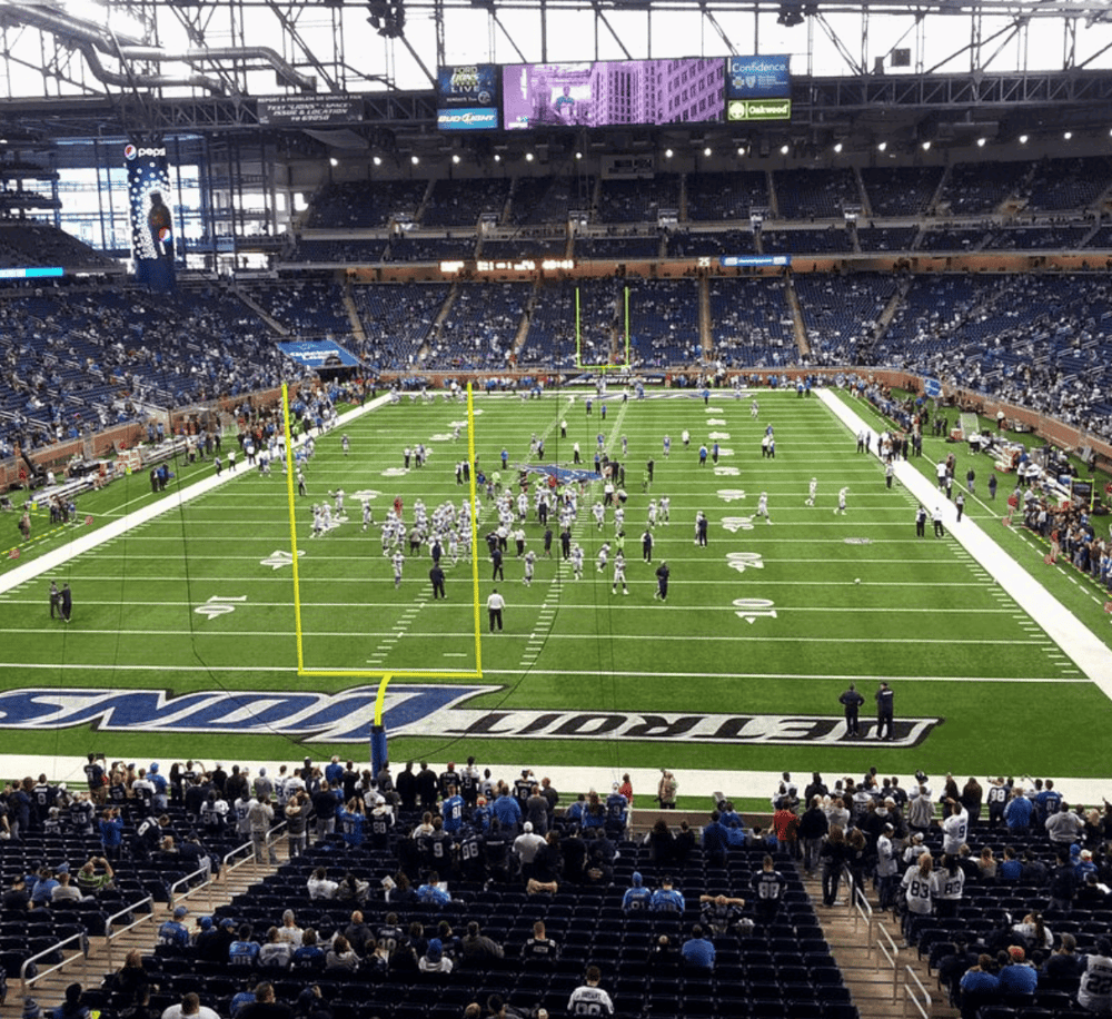 Full NFL stadium with players on field during a game, fans in seats, and digital scoreboards overhead.