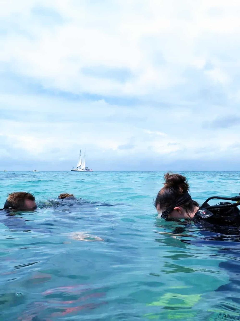 Scuba divers exploring clear blue ocean waters, vibrant marine life, and sailing boats in the background.
