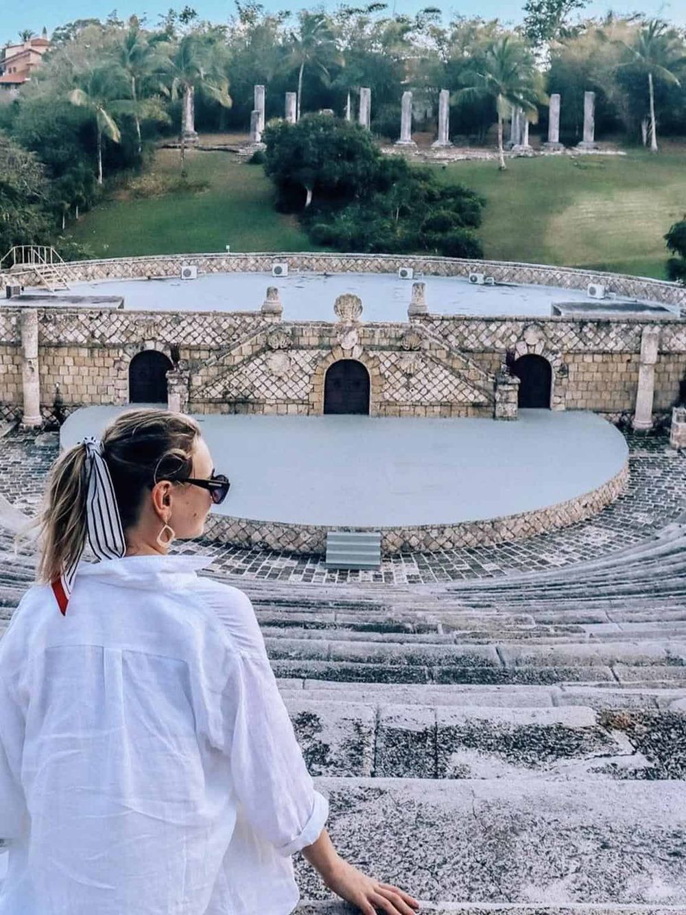 Ancient amphitheater with a woman overlooking the stage and lush greenery in the background.