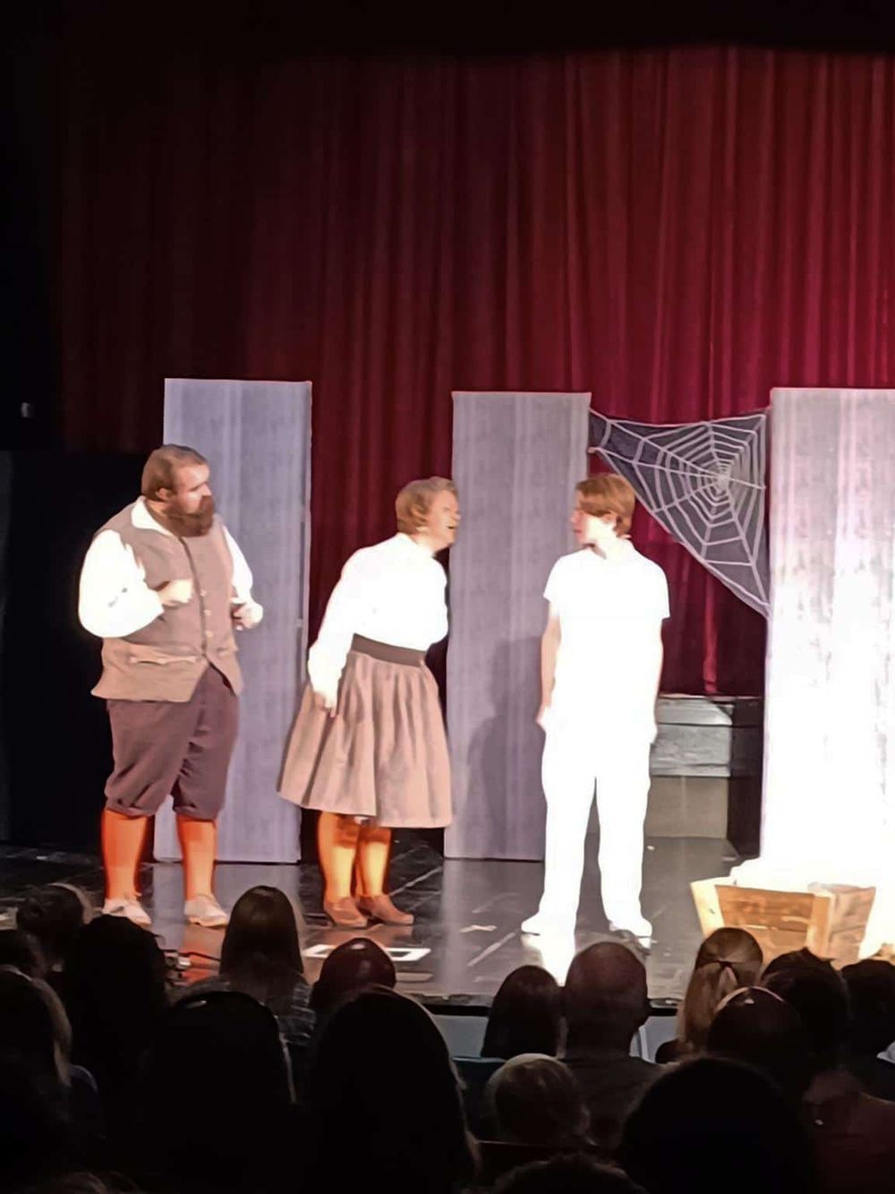 Dramatic scene from a theatrical play with actors in period costumes on stage with a spider web backdrop.