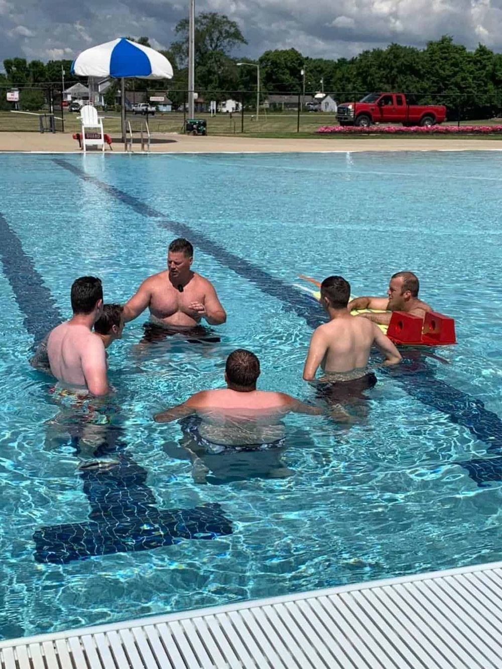 Adults participating in a swimming lesson at an outdoor pool with clear blue water, supervision, and safety equipment.