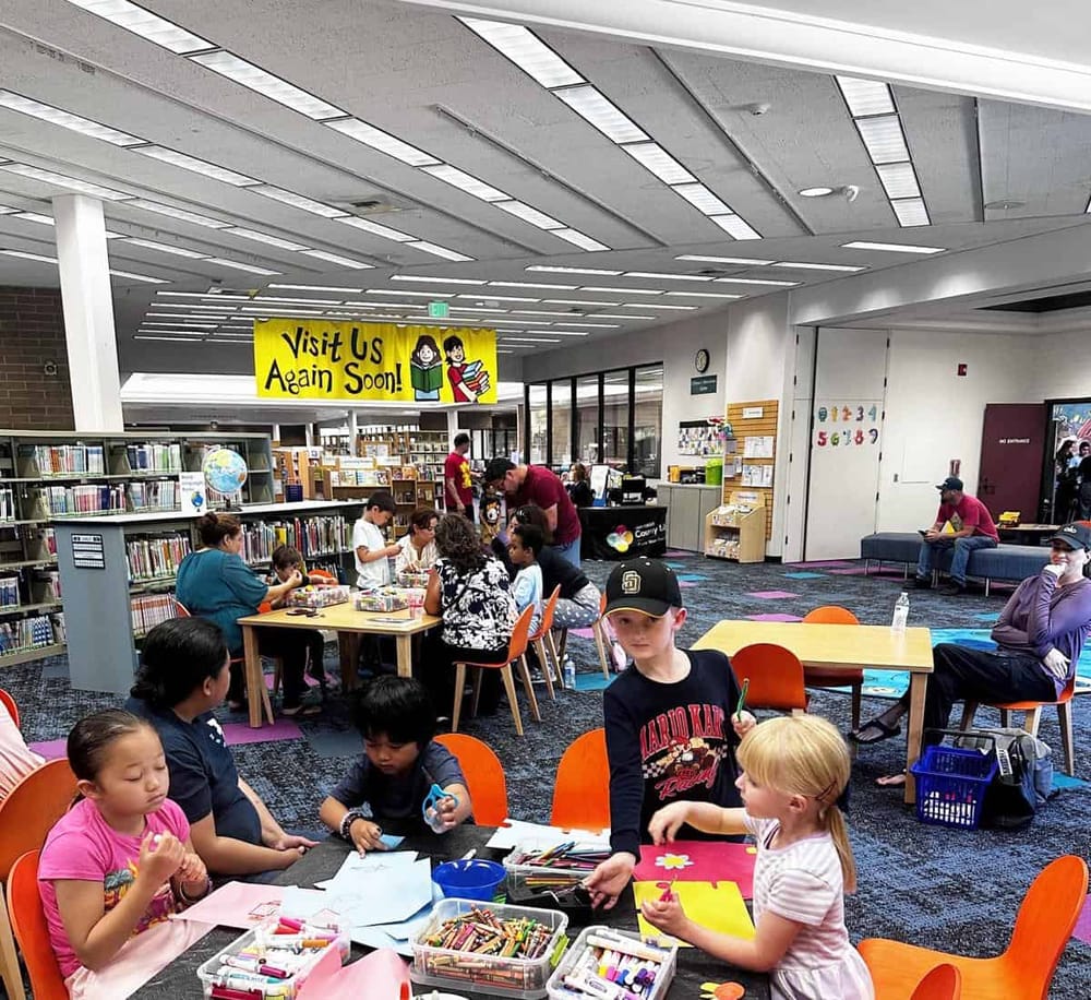 Colorful children's library activity room with kids engaged in arts and crafts, shelves of books, and a welcoming atmosphere.