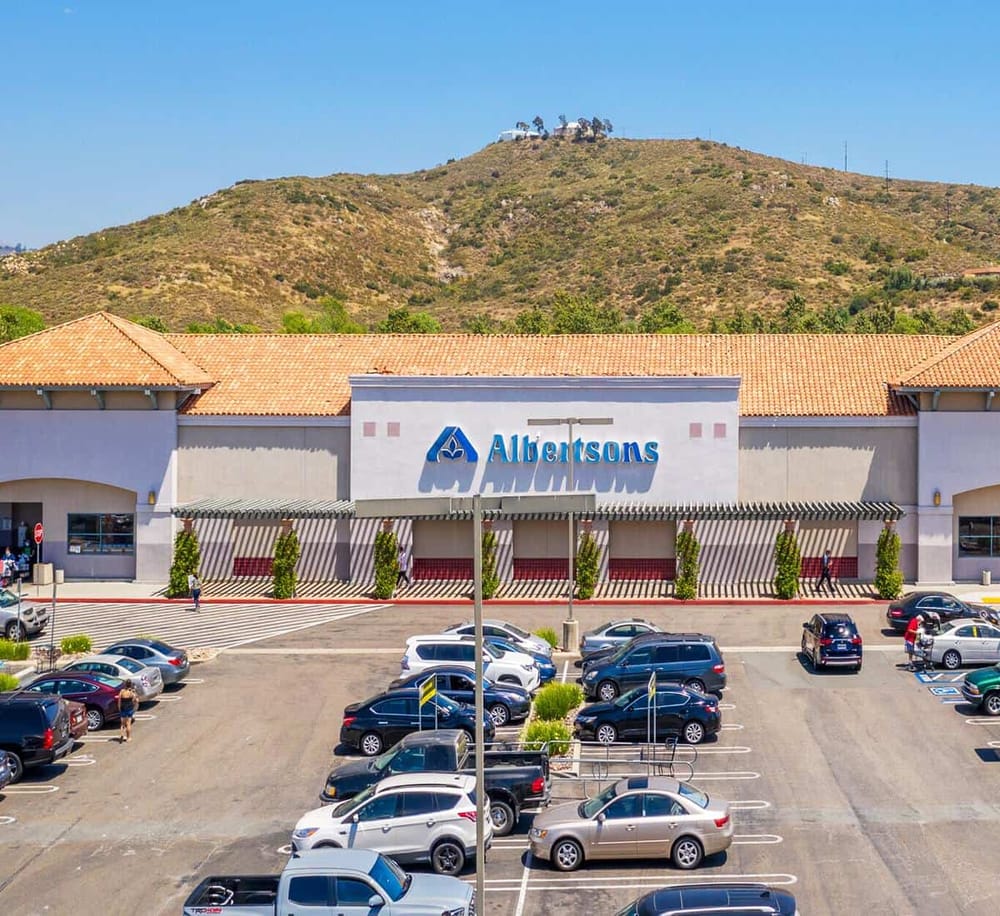 Albertsons grocery store exterior in shopping center, parking lot, with mountain backdrop.