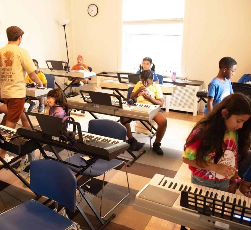 Children learning music in a classroom with keyboards and a teacher guiding.