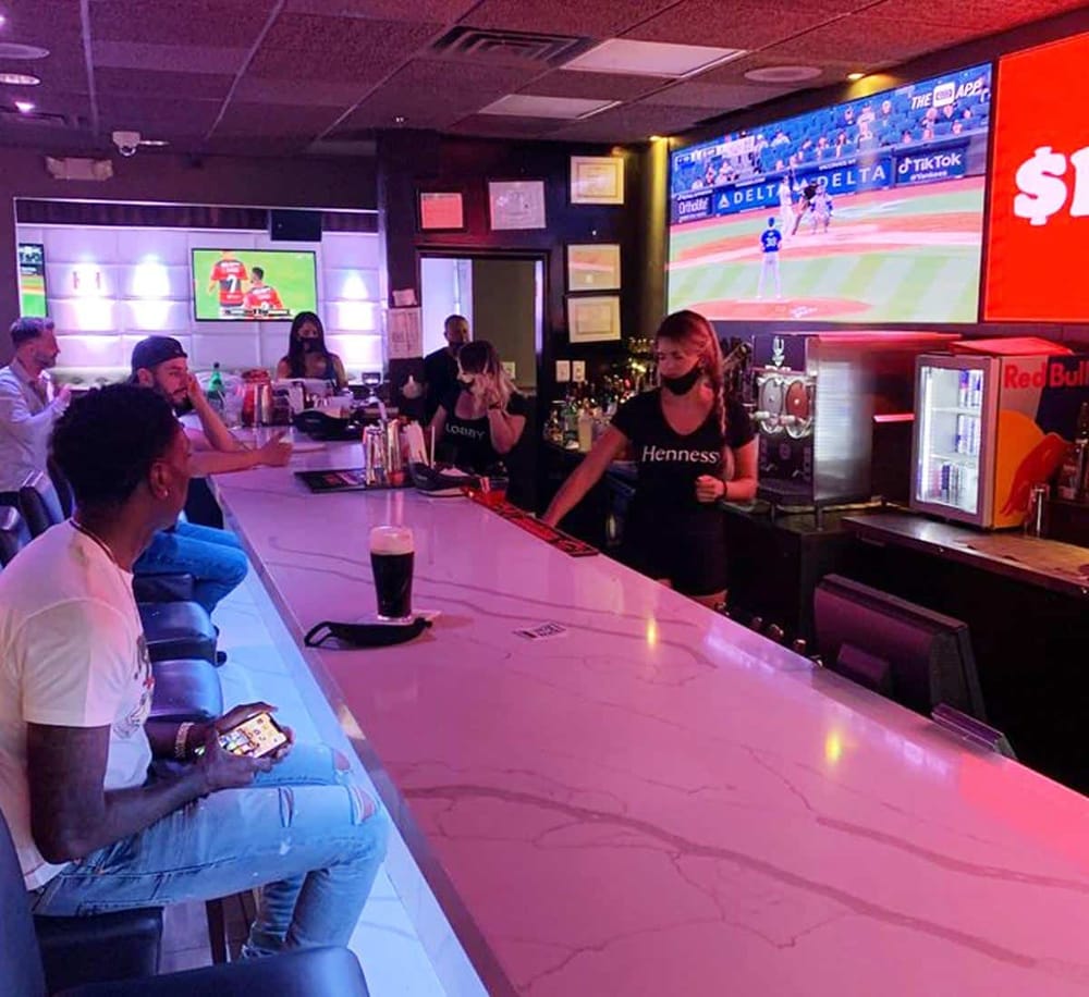 Relaxed sports bar with large TV screens showing baseball game, bar counter with drinks, and customers enjoying watching sports.