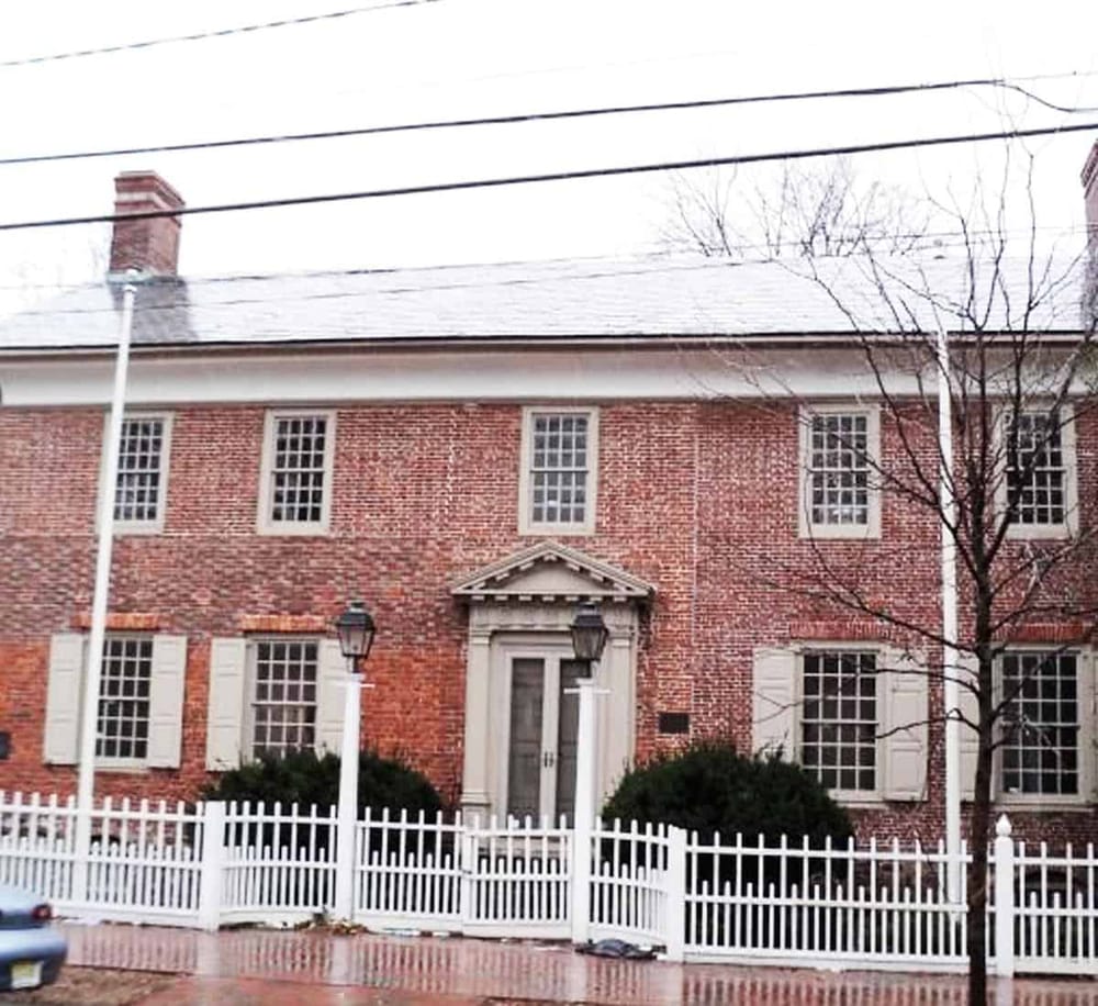 Brick historic house with white fencing and trees, located in a neighborhood setting.