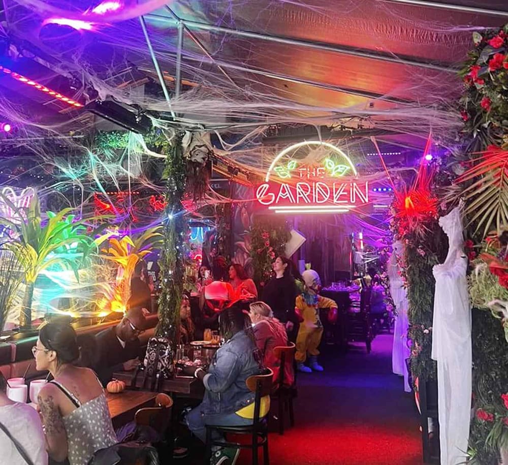 Neon lit sign reading "The Garden" with lively party scene and Halloween decorations.