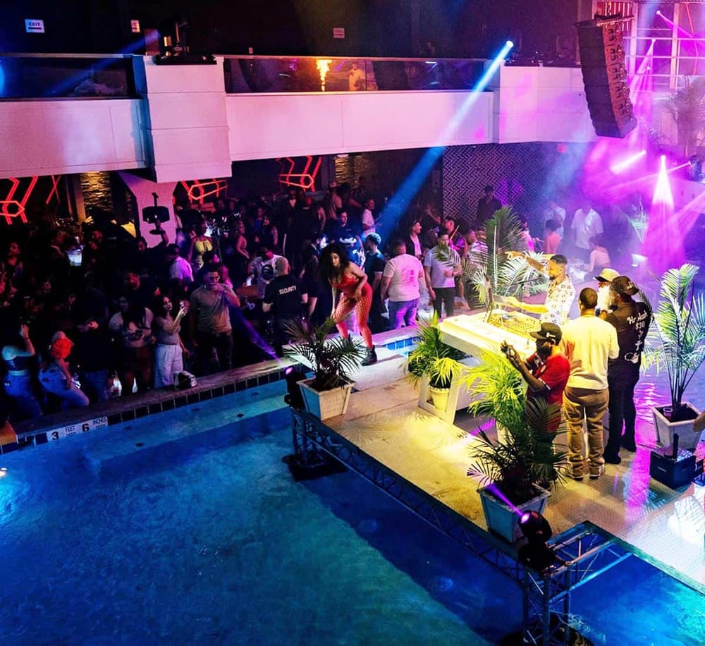 Bright nightclub with dancing crowd and DJ, lively atmosphere with colorful lights and tropical plants.