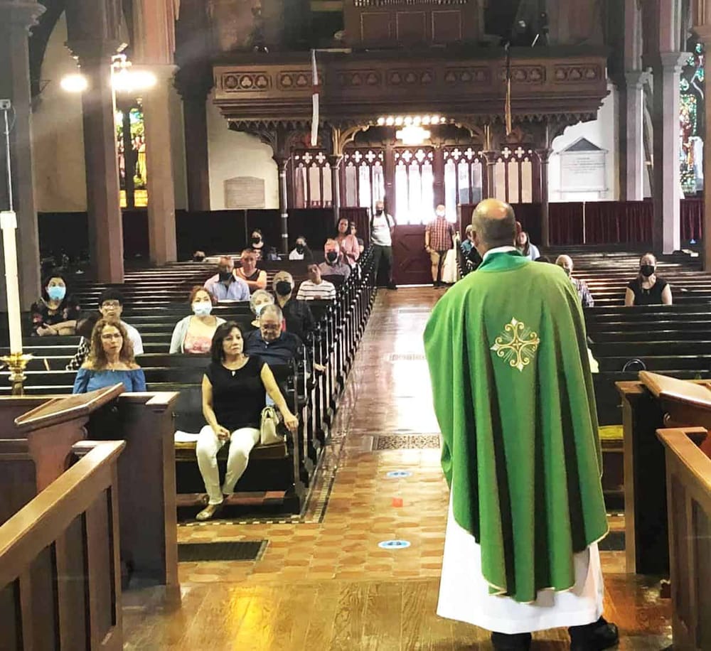 People attending church service inside a historic cathedral or church, with a priest in green robes leading the congregation.