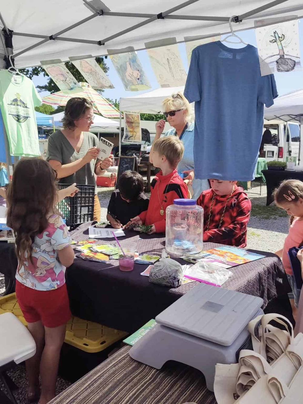 Colorful outdoor farmer's market stall with children and adults participating in a botanical activity.
