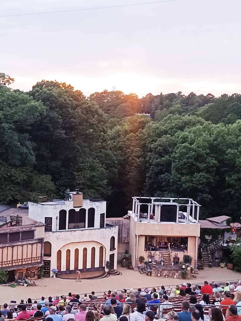 Theatre performance at sunset with audience in an outdoor amphitheater surrounded by lush trees.