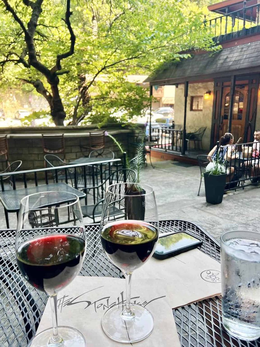 Cozy outdoor patio at a restaurant with wine glasses, lush green trees, and a charming building in the background.