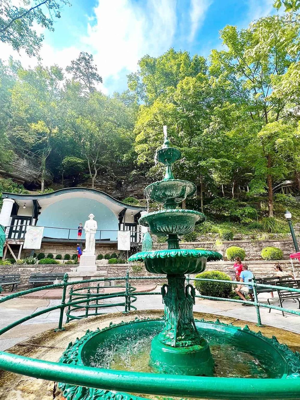 Bright green fountain at a park with lush trees in the background, featuring a sculpture and outdoor amphitheater, perfect for family outings and events.