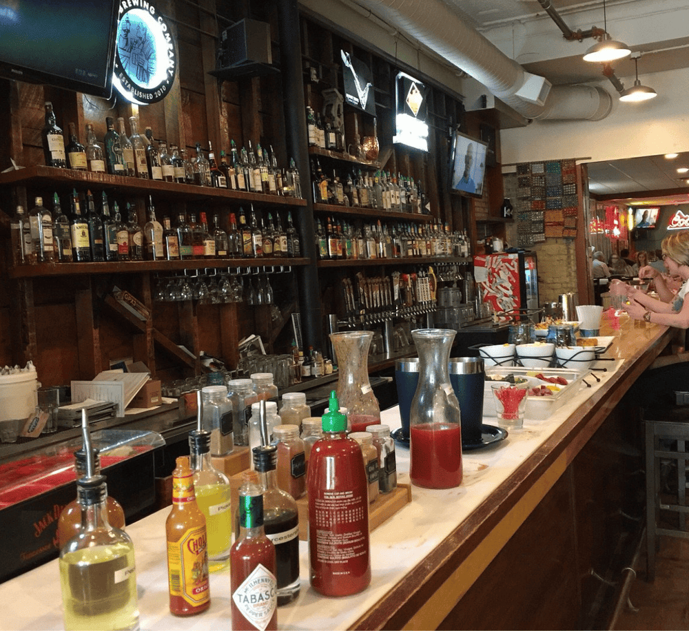 Clinking bar with liquor bottles, condiments, and customers enjoying drinks at Quest for Directions.