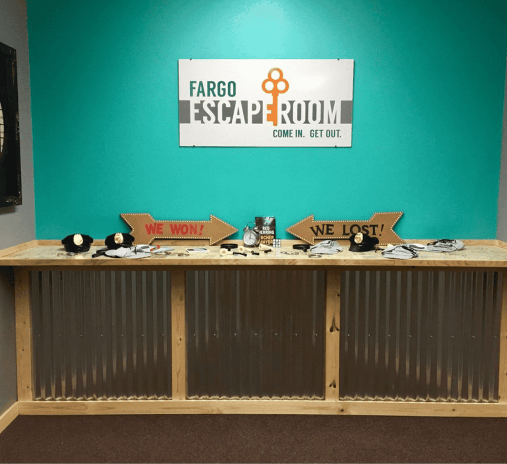 Escape room reception desk with signs indicating wins and losses, promotional items, and a QuestForDirections branding.