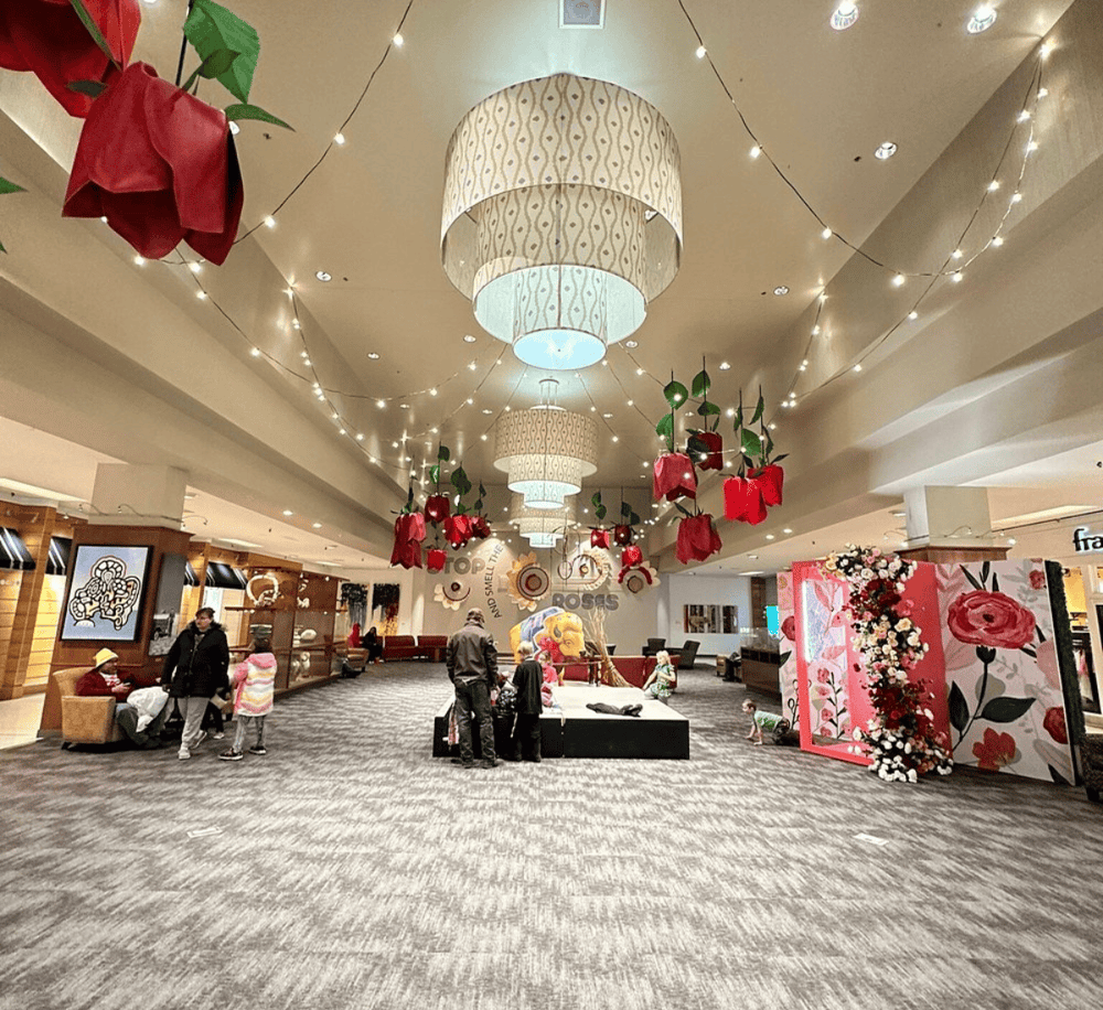 Bright indoor mall atrium decorated with large red roses and floral art displays, featuring festive lighting and modern decor.