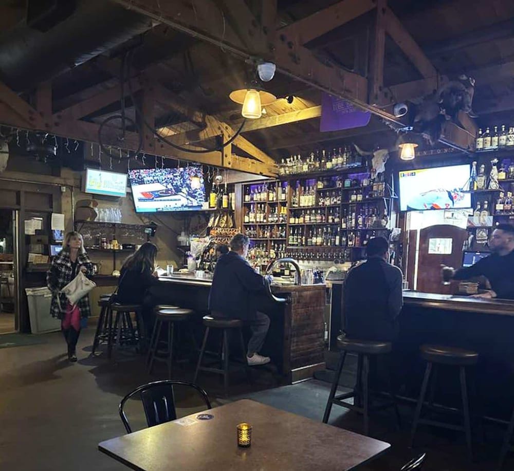 A cozy bar interior with wooden accents, multiple screens, and patrons enjoying drinks in a rustic setting.