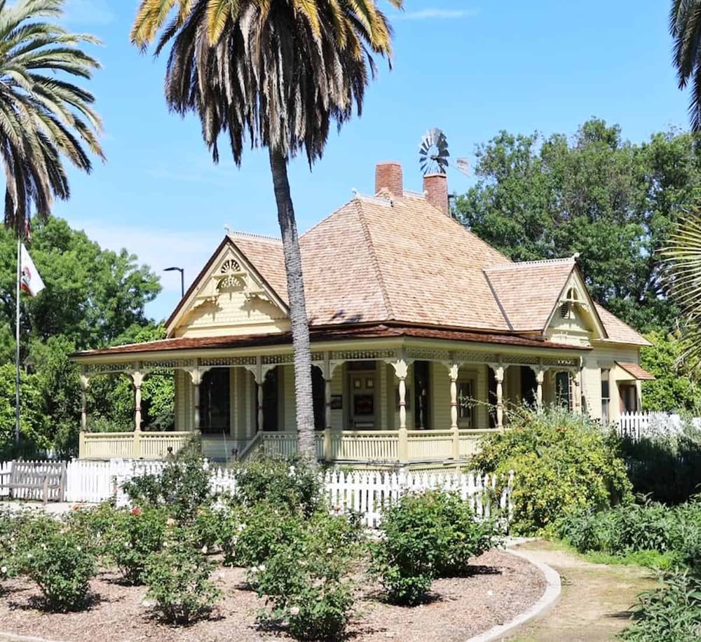 Victorian house with a wraparound porch, surrounded by lush greenery and tall palm trees, in a peaceful neighborhood.