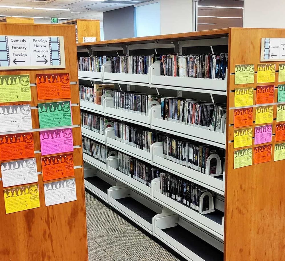 Aisle of DVDs and movies in a library or media store with colorful summer reading posters.