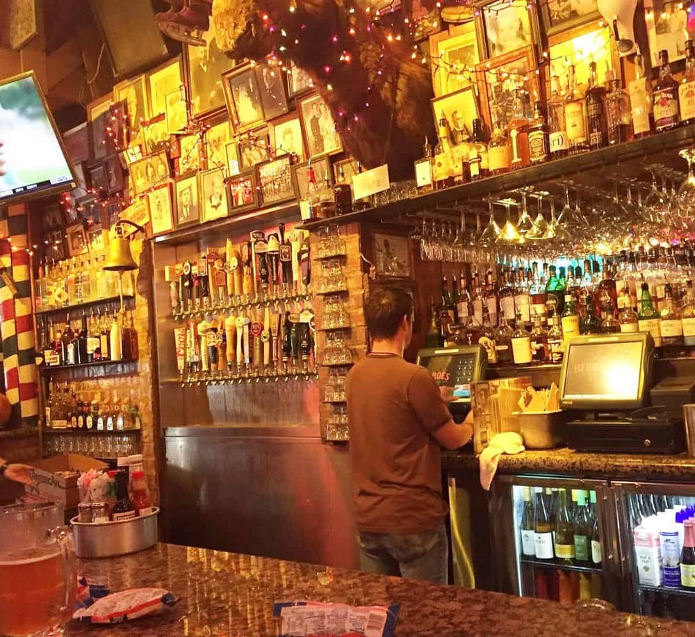 Colorful bar scene with an employee at a point-of-sale system, surrounded by liquor bottles and framed photos, warm lighting setting.