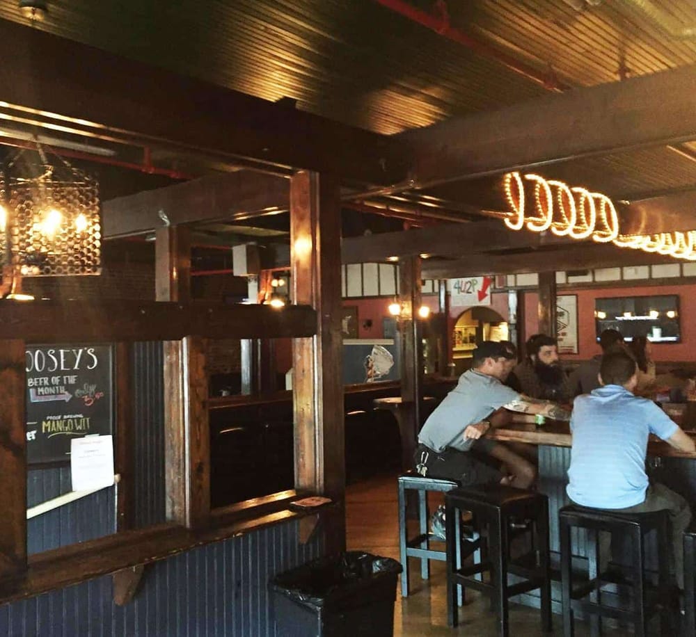A cozy bar interior with patrons sitting at high tables, warm lighting, and rustic wooden accents.