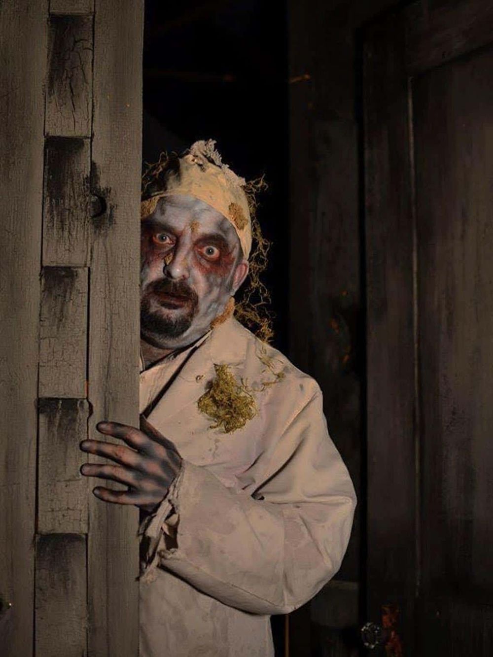Eerie haunted house actor peeking through wooden door with creepy makeup and costume for Halloween or horror theme.