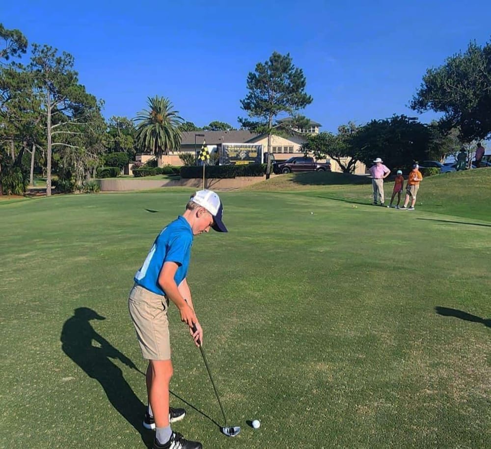 Boy golfing on a lush green golf course with a flag, trees, and clubhouse in the background.