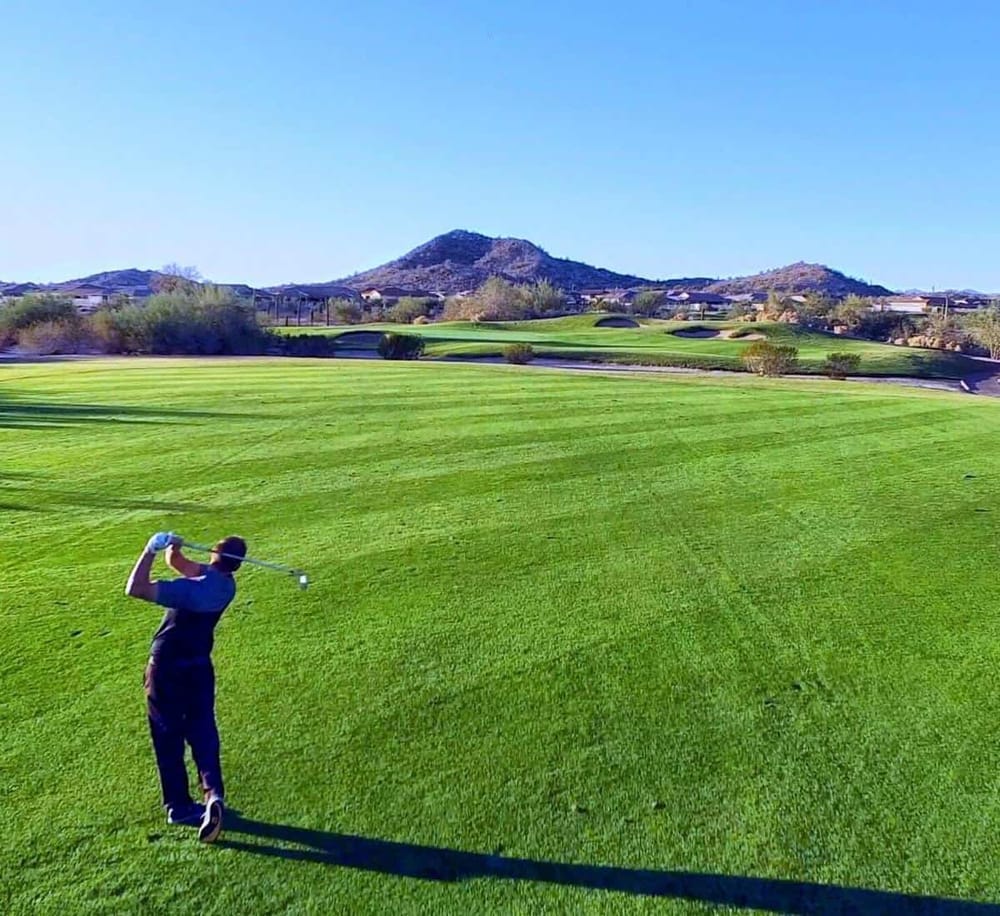 Aerial view of a golfer playing on a lush green golf course with mountain backdrop and clear blue sky.