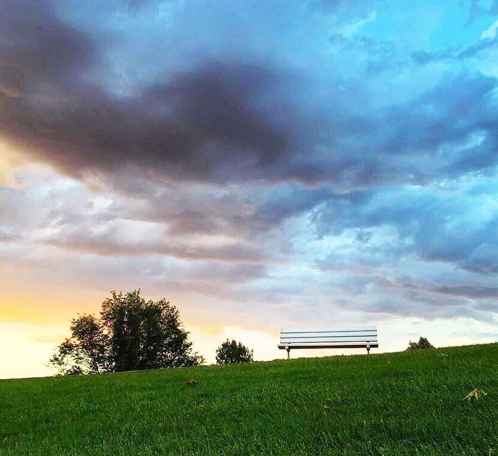 Empty park bench at sunset with dramatic cloudy sky, peaceful outdoor scenery, ideal for reflection and relaxation.